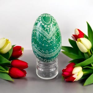 Op de afbeelding: Een groen paasei met een wit kantmotief wordt getoond op een transparante glazen standaard. Het ei wordt omringd door een boeket rode en witte tulpen.