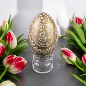 Peut inclure: Un œuf doré avec un motif floral blanc est présenté sur un support en verre transparent. L'œuf est entouré de tulipes roses et blanches.