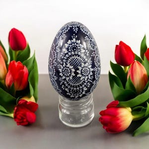 May include: A blue and white decorated Easter egg with a floral design sits on a clear glass stand. The egg is surrounded by red tulips.