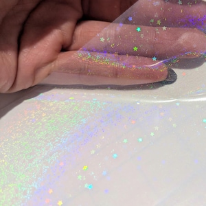 Puede incluir: Una lámina de material transparente iridiscente con una dispersión de diminutas lentejuelas de colores en forma de estrella. El material está sujeto por una mano, revelando su flexibilidad y propiedades de reflexión de la luz. El fondo es una superficie blanca lisa.