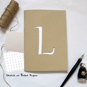 Puede incluir: Un cuaderno de papel kraft marrón con una letra "L" recortada en la portada. El texto "Sketch or Bullet Paper" está impreso en la parte inferior. También hay un rollo de cuerda, muestras de papel y una pluma estilográfica negra.