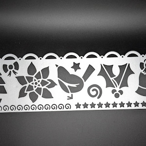 Puede incluir: Un esténcil de metal plateado con diseños recortados con temática navideña. Los diseños incluyen un árbol de Navidad, un lazo, una flor de pascua, un pájaro, una estrella, un remolino, acebo y una campana.