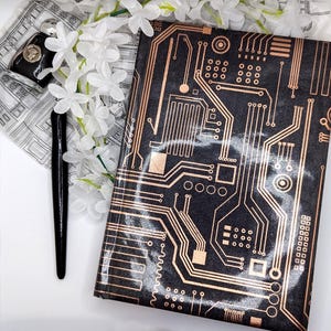 Puede incluir: Cubierta de cuaderno negra con un diseño de placa de circuito de color cobre. Un bolígrafo negro y un pequeño estuche negro con un emblema plateado descansan sobre una tela estampada. Flores blancas dispuestas cerca del cuaderno.