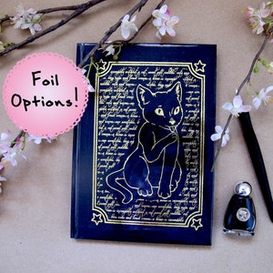 Puede incluir: Un diario azul oscuro con una ilustración de gato y texto en lámina dorada. Junto a él hay un tintero negro y una pluma. La imagen también incluye un círculo rosa con el texto "Foil Options!" y elementos florales.