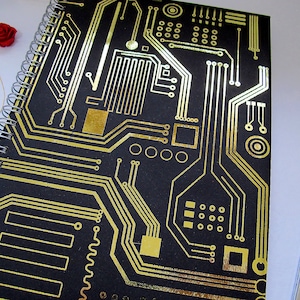 Pode incluir: Um caderno preto com design de placa de circuito dourado. A capa apresenta um padrão detalhado de linhas, quadrados e círculos dourados, que se assemelham aos componentes internos de um chip de computador. O design é dourado metálico sobre um fundo preto.