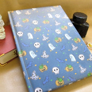 May include: A hardcover journal with a blue cover featuring a Halloween-themed pattern of pumpkins, ghosts, bats, witch hats, and skulls. A red book, a skull-shaped bottle, an ink bottle, and a glass pen are also visible.
