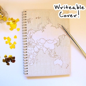 May include: A spiral-bound notebook with a world map design on the cover. The cover is light gray with a detailed outline of the continents in a gold color. A pen rests on the notebook, and the text "Writeable Cover!" is visible. Gold confetti is scattered around.