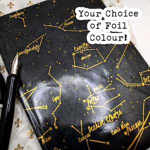 Puede incluir: Un diario negro con diseños de constelaciones doradas y texto. Una pluma estilográfica negra y un tintero están junto al diario. El texto "Your Choice of Foil Colour!" es visible.