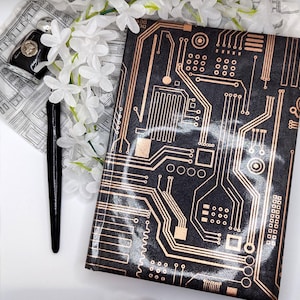 May include: Black notebook cover featuring a copper-coloured circuit board design. A black pen and a small black case with a silver emblem are next to the notebook. White flowers and a patterned scarf are also in the image.