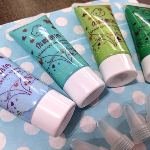 Puede incluir: Cuatro tubos de crema de simulación en azul, turquesa, verde claro y verde oscuro, cada uno con una tapa blanca. Los tubos tienen texto chino y diseños decorativos. También se ven varias puntas aplicadoras de plástico transparente.