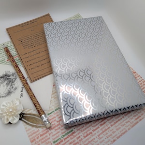 May include: A selection of vintage items, including a silver-patterned book, a pencil, a flower pin, and aged paper ephemera. The items are arranged on a white surface, creating a nostalgic aesthetic. The book has a scale pattern.