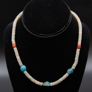 Puede incluir: Un collar artesanal con una cadena de cuentas circulares planas de color blanquecino. El collar está acentuado con vibrantes cuentas turquesas y rojas. Se muestra sobre un maniquí negro, mostrando su diseño y artesanía.
