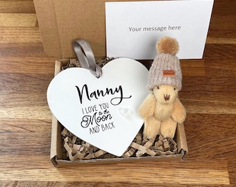 Könnte beinhalten: Weiße herzförmige Plakette mit dem Text "Nanny I love you to the moon and back" und einem kleinen Teddybär mit brauner Strickmütze in einer braunen Pappbox mit zerknülltem Papier.
