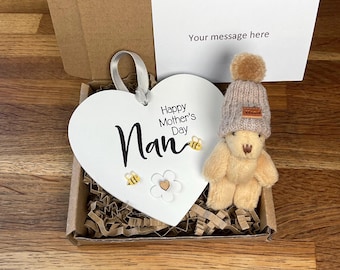 Könnte beinhalten: Ein weißes herzförmiges Holzschild mit dem Text "Happy Mother's Day Nan" und zwei kleinen gelben Bienen. Ein kleiner beiger Teddybär mit einer braunen Strickmütze ist in der Schachtel enthalten.