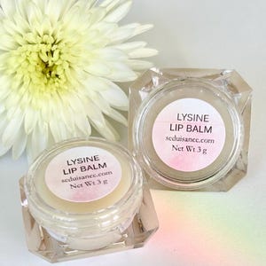 Può includere: Due barattoli di vetro trasparente di balsamo per labbra con etichette bianche che recitano "LYSINE LIP BALM" e "seduisancc.com" con un peso netto di 3 grammi.
