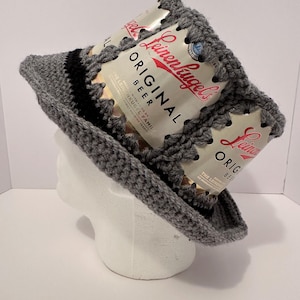 May include: A gray crocheted hat with a black band. The hat is decorated with two empty beer cans, one on top of the other. The cans are from Leinenkugel's Original Beer.