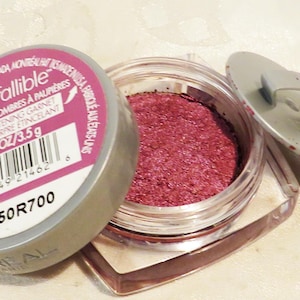 May include: An open jar of L'Oréal Infallible eye shadow in the shade 557 Glistening Garnet. The jar is clear with a silver lid and contains a sparkling, deep pink powder. The label is pink and silver.