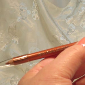 May include: A copper-colored eyeliner pencil with a clear plastic cap. The pencil is held in a hand, and the background is a light, patterned fabric.