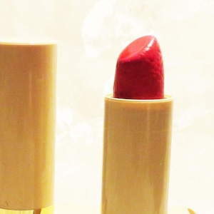 May include: A vintage Avon lipstick in a beige tube with a gold band. The lipstick is a vibrant red color and is partially extended. The lipstick is next to its original box, which has the words "AVON SATIN SMOOTH LIPSTICK" printed on it.