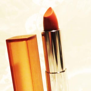 May include: A lipstick with a bright orange-red hue, displayed with its cap. The lipstick tube is silver with a geometric design, and the cap is a translucent orange. The product is set against a soft, off-white background.
