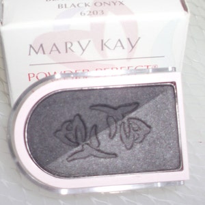 May include: A Mary Kay Powder Perfect eyeshadow compact in Blackest Black/Black Onyx. The compact is a light pink with a dark gray eyeshadow divided into two shades. The eyeshadow has a floral design embossed on the surface.