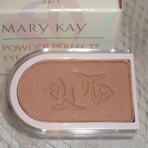 May include: A Mary Kay Powder Perfect eye shadow compact in the color "Seashell." The compact is a light pink with a clear plastic top. The eye shadow is a light brown color with a floral design embossed on the surface. The box is off-white with the Mary Kay logo.