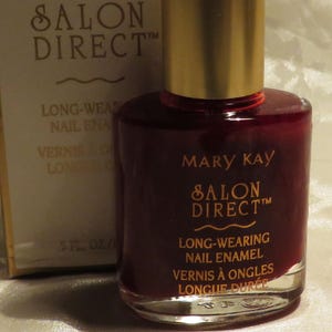 May include: A bottle of Mary Kay Salon Direct long-wearing nail enamel in a deep red shade. The bottle has a gold cap and is next to its matching box. The box and bottle both feature the Mary Kay logo and the words 'Nail Enamel'.