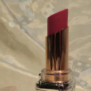 May include: Open lipstick tube with a bright pink lipstick bullet. The lipstick has a gold-colored metal casing and a clear, geometric-patterned plastic base. The cap is off and next to the lipstick. The background is a light, patterned fabric.