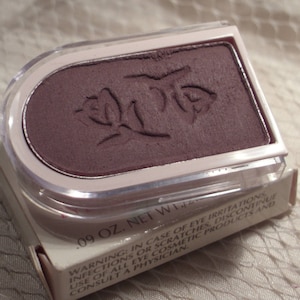 May include: A plum-colored eyeshadow compact with a clear lid and a white frame. The eyeshadow has a floral design embossed on its surface. The packaging includes a box with text that reads, ".09 OZ. NET WT."