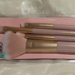 May include: A set of makeup brushes in a clear plastic package. The brushes have light pink handles with gold-colored ferrules and light pink bristles. The package is on a pink background with the words "Studio Lux" in teal.