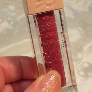 May include: A Maybelline lip gloss tube with a light peach cap and a clear body. The tube contains a red lip gloss. The word "LIFTER" is embossed on the cap. The words "MAYBELLINE NEW YORK" are printed on the tube.
