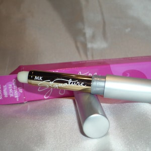May include: A silver lip liner pencil with the word "Signature" in cursive script. The pencil is partially extended, revealing the lip liner. A pink box with the words "Lip Outliner Pencil" is in the background.
