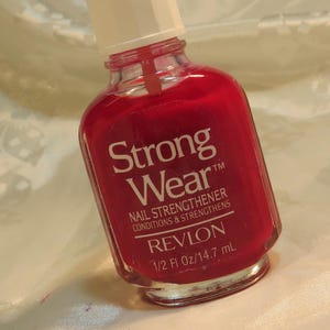 May include: A bottle of Revlon Strong Wear nail strengthener in a vibrant red hue. The bottle has a white cap and features the product name and details, including "1/2 Fl Oz/14.7 mL". The nail polish is designed to condition and strengthen nails.