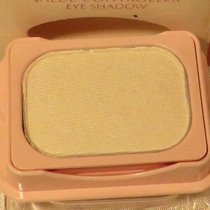 May include: A light pink eye shadow compact with a pale yellow eye shadow. The compact has a rectangular shape with rounded corners and a clear plastic base. The eye shadow has a textured surface.