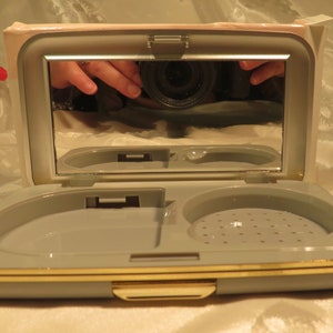May include: Open, vintage compact case in a light gray color with a gold-colored trim. The compact has a mirror on the inside of the lid and a compartment for powder and other cosmetics. The case is rectangular and has a clasp closure.