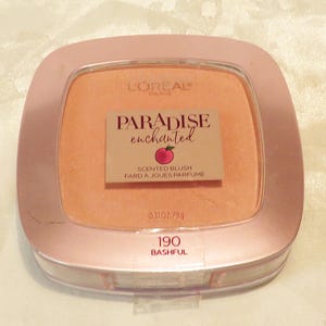 May include: A peach-toned blush compact with a rose gold casing. The compact is branded with "L'OREAL PARIS" and "PARADISE enchanted". The product is labelled "SCENTED BLUSH FARD A JOUES PARFUME" and "190 BASHFUL". Contains 9g of product.