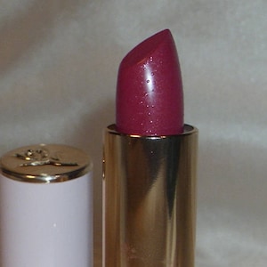 May include: Close-up of a lipstick with a deep berry color. The lipstick is in a gold-colored tube with a white base and a gold cap. The lipstick has a shiny finish and appears to be new.