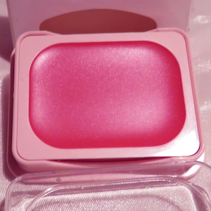 May include: A pink lip product in a rectangular compact. The product has a smooth, shiny surface and is encased in a light pink plastic container with a clear lid. The background is a soft, light pink.