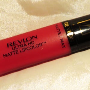May include: A red Revlon Ultra HD Matte Lipcolor tube with a black cap and gold accents. The tube is matte and has the Revlon logo and product information printed on it. The background is a cream-colored fabric.