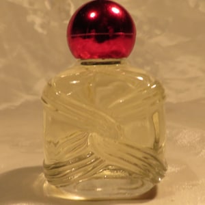 Peut inclure: Un flacon de parfum en verre transparent avec un bouchon rouge. Le flacon a un design texturé et est posé sur une surface blanche.