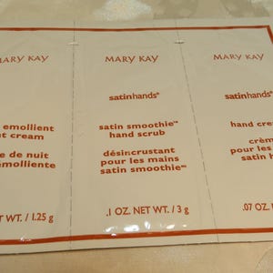 May include: A white packet with three sections, each labeled "MARY KAY" and containing product information. The sections list "extra emollient night cream", "satin smoothie hand scrub", and "hand cream". Measurements are provided in ounces and grams.