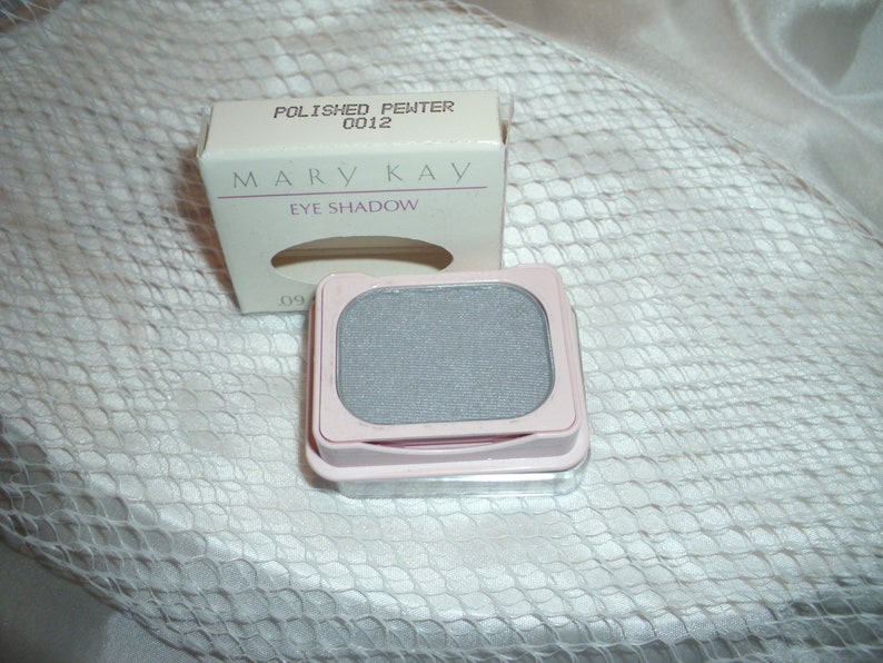 May include: A small, square, silver eye shadow compact with a pink lid. The compact is labeled 'Mary Kay Eye Shadow' and 'Polished Pewter 0012'.