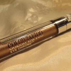 May include: A tube of Maybelline Color Tattoo Eye Chrome eyeshadow in a bronze color. The tube is clear with a silver cap and the product name is printed in brown. The eyeshadow is held by a person.