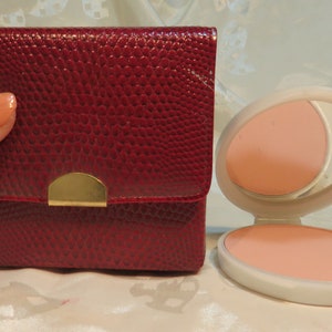 Puede incluir: Un estuche compacto de imitación de cuero rojo con cierre dorado y un espejo y un estuche de polvo a juego. El estuche de polvo está abierto y el polvo es de color rosa claro.
