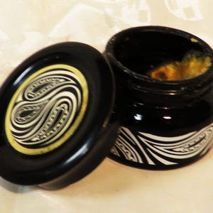 May include: A small black jar with a matching lid, both featuring a white and yellow swirling graphic pattern. The jar is open, revealing a textured, yellowish substance inside. The design consists of curved lines and shapes on the exterior.