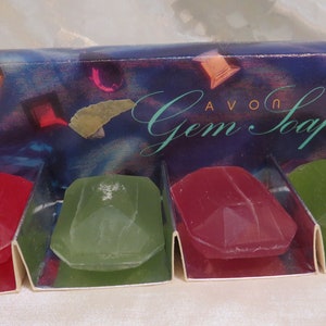 May include: Four jewel-shaped soaps in a clear plastic holder. The soaps are red, green, red, and green. The box reads "Avon Gem Soaps".
