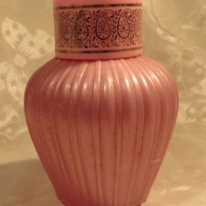 May include: A pink glass bottle with a gold and white paisley design on the lid. The bottle has a ribbed design and is empty.
