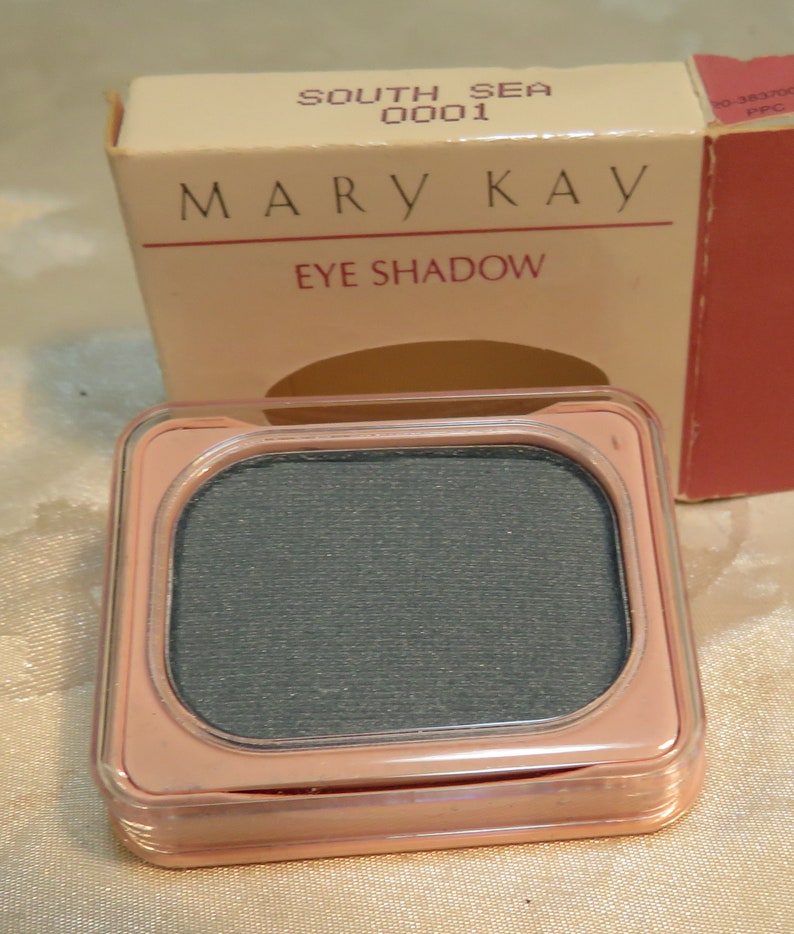 Peut inclure: Un fard &agrave; paupi&egrave;res Mary Kay dans un bo&icirc;tier p&ecirc;che clair, contenant un fard &agrave; paupi&egrave;res gris fonc&eacute;. La bo&icirc;te du produit est visible en arri&egrave;re-plan, avec les mots "Mary Kay" et "Eye Shadow" imprim&eacute;s dessus.