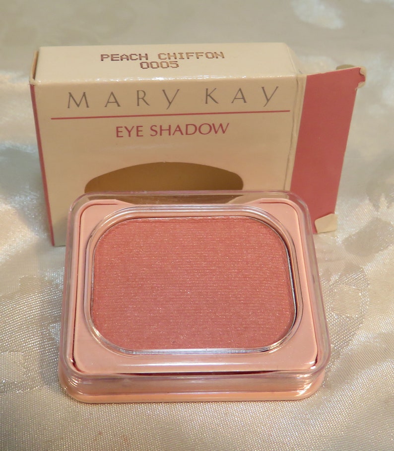 May include: Mary Kay Peach Chiffon Eye Shadow in a translucent pink compact. The compact has a square shape with rounded corners. The eyeshadow is a light peachy-pink shade. The packaging is light beige with dark brown text that reads 'Mary Kay' and 'Eye Shadow.' The lid also indicates the shade name 'Peach Chiffon' and the number '0005'.
