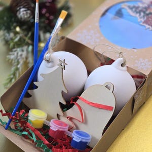 May include: A craft kit for decorating Christmas baubles. The kit includes two white ceramic baubles, two wooden baubles, three paint pots, two paintbrushes, and red and green shredded paper.